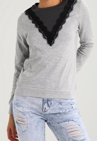Sudadera gris con detalles de encaje negro en el escote en V, mangas largas y puños acanalados, combinada con vaqueros claros y desgastados.