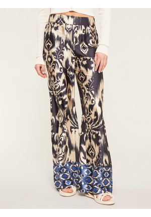Kobieta nosząca spodnie z wysokim stanem i szerokimi nogawkami w beżowo-czarne abstrakcyjne wzory oraz niebieskie wzorzyste mankiety, zestawione z białymi sandałami.