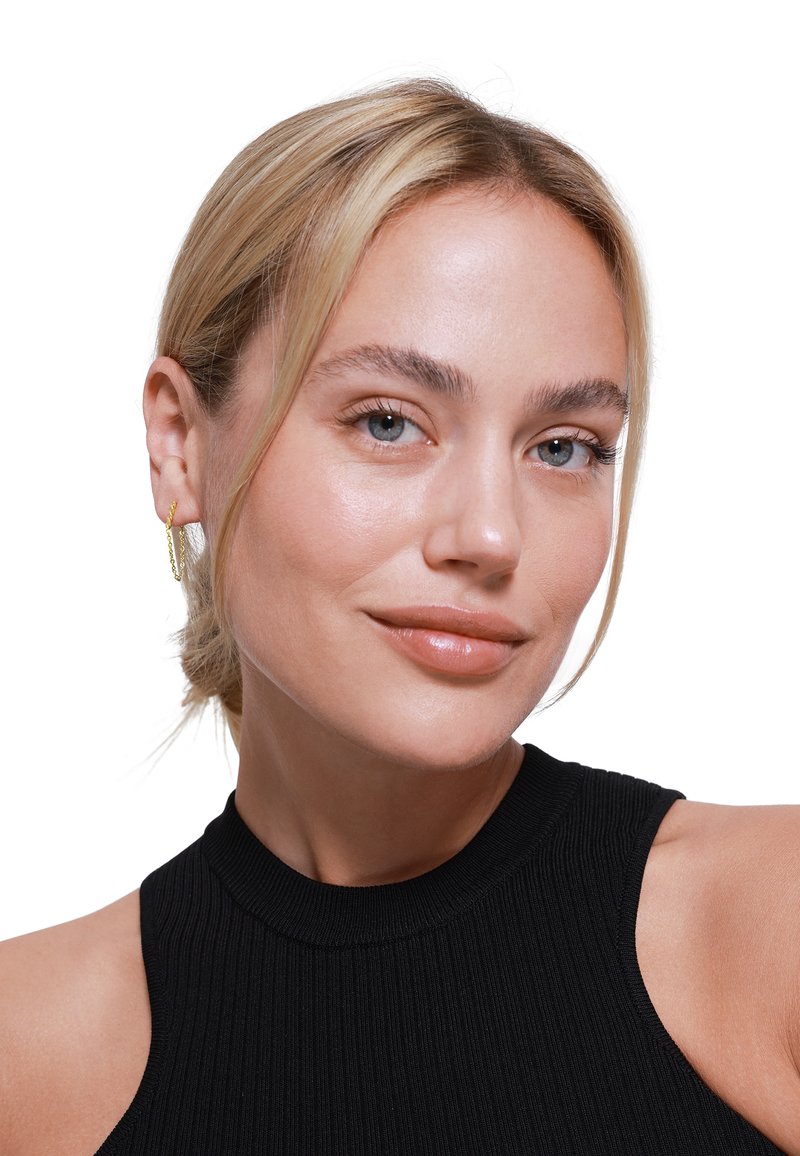 Boucles d'oreilles créoles torsadées en orportées par une personne avec des cheveux blonds clairs, une peau lisse et un haut noir ajusté à côtes, sur fond blanc.