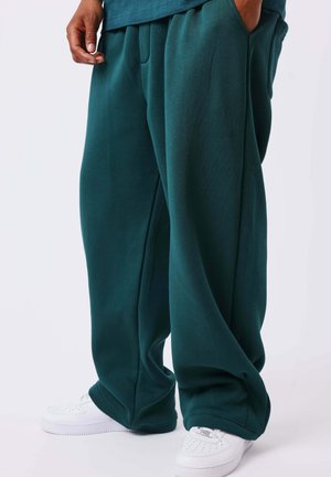 Personne portant un pantalon vert foncé à jambes larges, mains dans les poches et baskets blanches sur un fond clair.