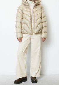 Beige péřová bunda se zipem vpředu a kapucí, zdobená šikmým prošíváním. V kombinaci se širokými světle krémovými kordovými kalhotami a tmavě hnědými botami.