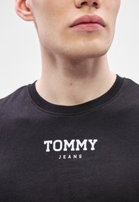 T-shirt preta de algodão com um logótipo branco impresso que diz "TOMMY" acima de "JEANS". Design com gola redonda e textura suave.