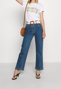 Camiseta blanca con gráfico floral, jeans de mezclilla azul de cintura alta, cinturón tejido beige y sandalias de tacón negras. Incluye un bolso redondo marrón.