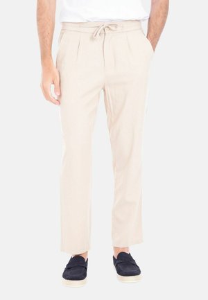 Uomo che indossa pantaloni beige con coulisse, camicia bianca infilata dentro e scarpe slip-on nere con suole bianche, in piedi di fronte a chi guarda.