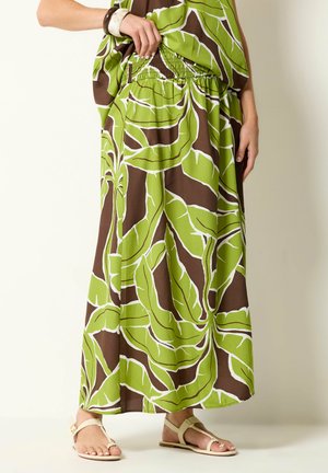 Femme portant une longue jupe à motif de feuilles vertes et marron avec une taille élastique et des sandales plates beiges, debout devant un fond uni.