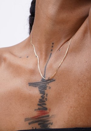 Gold chain necklace with a zigzag design, resting on bare skin, alongside intricate tattoos featuring black and red ink patterns.