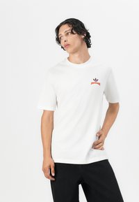 White cotton t-shirt with ribbed crew neck, short sleeves, and red Adidas logo on the left chest. Worn with black pants.
