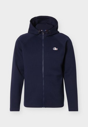 Μπλε ναυτικό φούτερ με φερμουάρ, εμπρόσθιες τσέπες, ρυθμιζόμενο σκουρόχρωμο κορδόνι στο κουκούλι και μικρό λευκό, μπλε και κόκκινο λογότυπο κροκοδείλου Lacoste στο στήθος.
