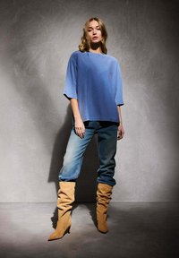 Chemise bleue surdimensionnée à manches courtes associée à un jean bleu clair et des bottines beige à talons décontractées, sur un fond gris texturé.