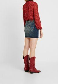 Donna che indossa una blusa rossa a fantasia, una mini gonna di jeans blu sbiadita e stivali alti fino al ginocchio in pelle rossa, in piedi su uno sfondo bianco semplice.
