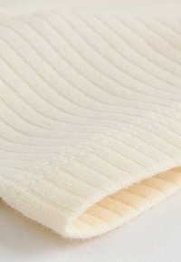 Cremfarbener gerippter Stoff mit einer weichen Textur, der feine vertikale Linien und eine gefaltete Kante zeigt, auf einem hellen Hintergrund liegend.