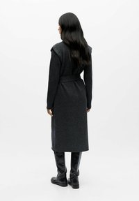 Cardigan lungo di un grigio scuro, con vita cinturata, maniche a coste e texture liscia. Indossato con stivali neri fino al ginocchio. Design minimalista, senza fantasie.