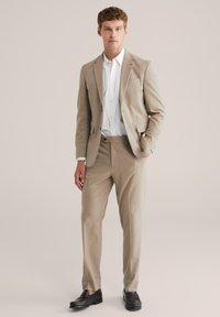 Jeune homme debout portant un costume beige, une chemise blanche et des mocassins marron foncé, contre un fond clair et uni.