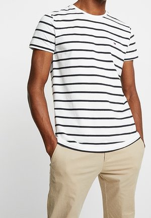 Mężczyzna w białej koszulce z krótkim rękawem w czarne poziome paski i beżowych spodniach, z rękami swobodnie w kieszeniach, na białym tle.