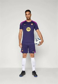 Barcelona FC fotbollskit i lila med rosa detaljer. Kort ärm, logga på bröstet, matchande shorts. Spelaren håller i en vit fotboll.