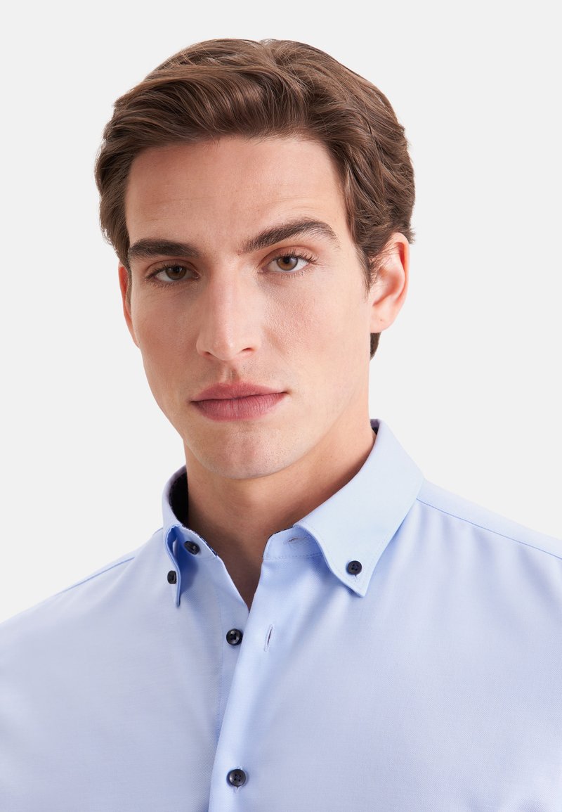 Jeune homme aux cheveux bruns coiffés, portant une chemise bleu clair boutonnée sur un fond blanc uni.