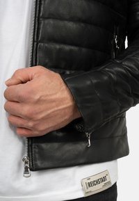 Zwarte leren jas met gestikt ontwerp, zilveren ritssluiting en ritszakken. Draag ik over een wit shirt met een label waarop 'REICHSTADT' staat.