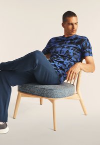 Camiseta de camuflaje azul de mangas cortas, combinada con pantalones azul marino. El modelo está sentado en una silla de madera con un cojín azul texturizado.