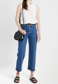 Biały top bez rękawów z koronkowymi detalami, niebieskie dżinsy z wysokim stanem, czarny pasek z ćwiekami, czarna torba na ramię i czarne klapki.