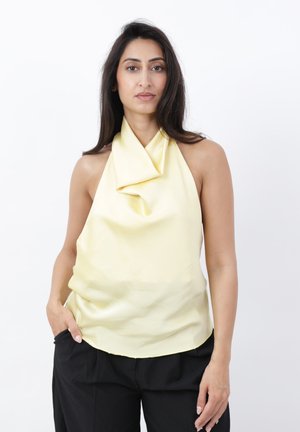 Femme portant un haut dos nu drapé jaune pâle et un pantalon noir, debout avec une main dans la poche sur fond blanc.