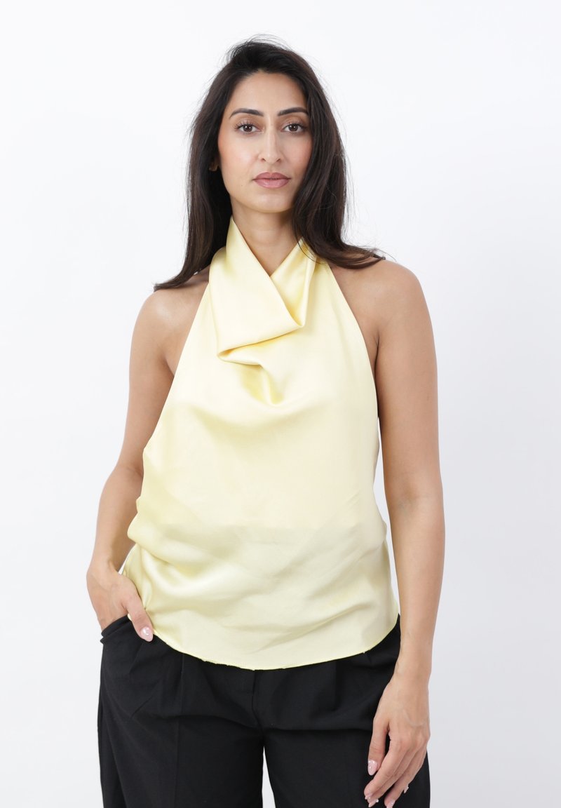 Mujer con top halter drapeado sin mangas de color amarillo pálido y pantalones negros, de pie con una mano en el bolsillo sobre fondo blanco.