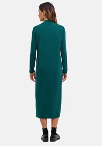 Robe longue ajustée en tissu vert foncé, avec un col et des manches longues. Longueur mi-mollet avec une texture lisse et des détails de couture subtils.