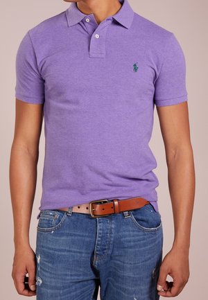 Lila Polo-Shirt mit kurzen Ärmeln aus strukturiertem Stoff, mit grünem Logo auf der Brust. Trägt man zu abgewetzten blauen Jeans und einem braunen Gürtel.