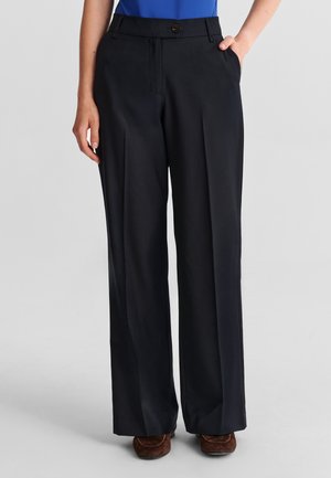 Femme portant un pantalon noir à jambes larges, un haut bleu et des chaussures marron, debout avec une main dans la poche sur un fond gris clair.