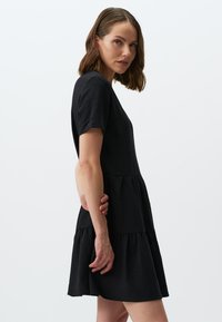 Zwarte katoenen jurk met korte mouwen, gelaagde zoom en een losse pasvorm, die een gladde textuur en minimalistisch ontwerp toont.