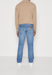 Modré denimové džíny rovného střihu, s hnědou koženou nášivkou na pase a zadních kapsách, nošeny s béžovým svetrem.