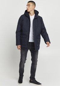Mørkeblå dunjakke med hette, med glidelås, to frontlommer og quiltet tekstur. Kombinert med gråjeans og svarte joggesko.