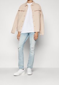 Ljust beige skjortjacka, vit t-shirt, ljusblå slitna jeans och vita sneakers. Enkelt mönster med fickor och avslappnad passform.