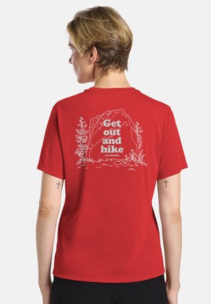 Persoon met kort haar die een rood T-shirt draagt met een wit ontwerp met natuurthema en de tekst "Get out and hike" op de rug, van achteren gezien.