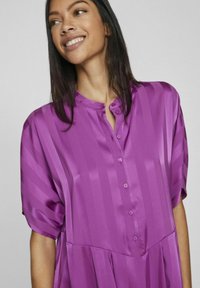 Blusa in satin viola con righe verticali, scollatura rotonda con bottoni e maniche corte. Il tessuto ha una texture liscia e lucida.
