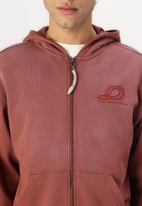 Kapišoninė zip-up džemperis raudonai rudos spalvos audinyje. Su priekinėmis kišenėmis, išsiuvinėtu logotipu ir tekstūruotu užtrauktuko traukimu su pintu šonu.