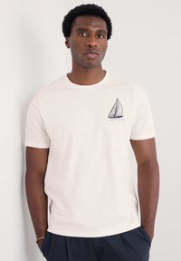 Camiseta de algodón blanca con cuello redondo, que presenta un gráfico de un velero azul en el lado izquierdo del pecho. Mangas cortas y un corte relajado.