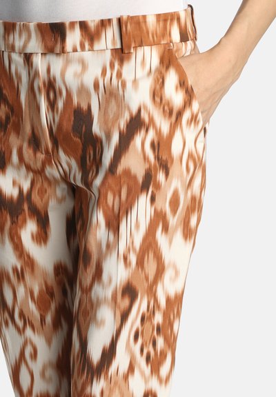 Pantalon avec un motif ikat marron et crème, doté d'une ceinture plate, de poches latérales et d'une texture de tissu lisse et légèrement brillante.