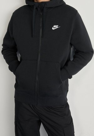 Czarna bluza z kapturem Nike na zamek z przednimi kieszeniami, logo na piersi, noszona przez osobę z rękami w kieszeniach na jednolitym tle.