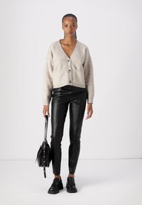 Lichtbeige cropped cardigan met een V-hals, knoopsluiting en zakken, gecombineerd met nauwsluitende zwarte leren broek en zwarte schoenen.