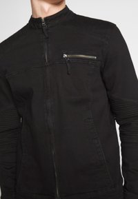 Veste en denim noire avec un col montant, une fermeture éclair à l'avant et une poche poitrine zippée. Présente des textures côtelées sur les manches.