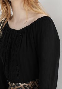 Mustaton, olkapäiden yli ulottuva pusero, jossa on pleatattu pinta ja pystysuorat virkattu yksityiskohdat. Pusero on yhdistetty ruskeansävyiseen, kuvioliseen hameeseen.