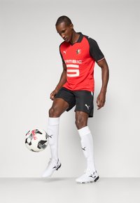 Un calciatore maschio in una maglietta rossa e nera e pantaloni neri bilancia un pallone su un piede, mostrando un'espressione concentrata.