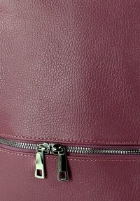 Een bordeauxleren tas met een gestructureerde afwerking, voorzien van een zilveren ritsluiting en rechthoekige ritsgrepen.