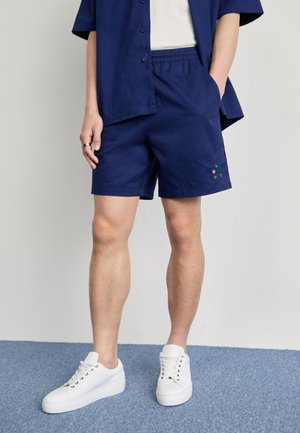 Person trägt marineblaue Shorts und passendes Hemd, weißes T-Shirt und weiße Sneaker, steht auf blauem Teppich mit den Händen in den Taschen.