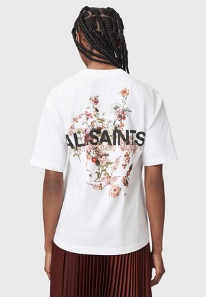 Person med flettet hår bærer en hvid t-shirt med blomsterdesign og teksten "ALLSAINTS" på ryggen, kombineret med en brun plisseret nederdel.