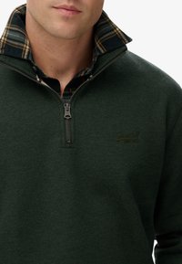 Pulóver de forro polar verde con cuello de cremallera y camisa a cuadros asomando por debajo. Tiene un cierre de cremallera y un sutil logo en el pecho.