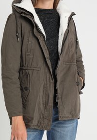 Chaqueta parka de color verde oliva con un forro de forro polar blanco esponjoso, capucha con cordón, bolsillos frontales y cierre de cremallera, llevada sobre una camisa oscura.