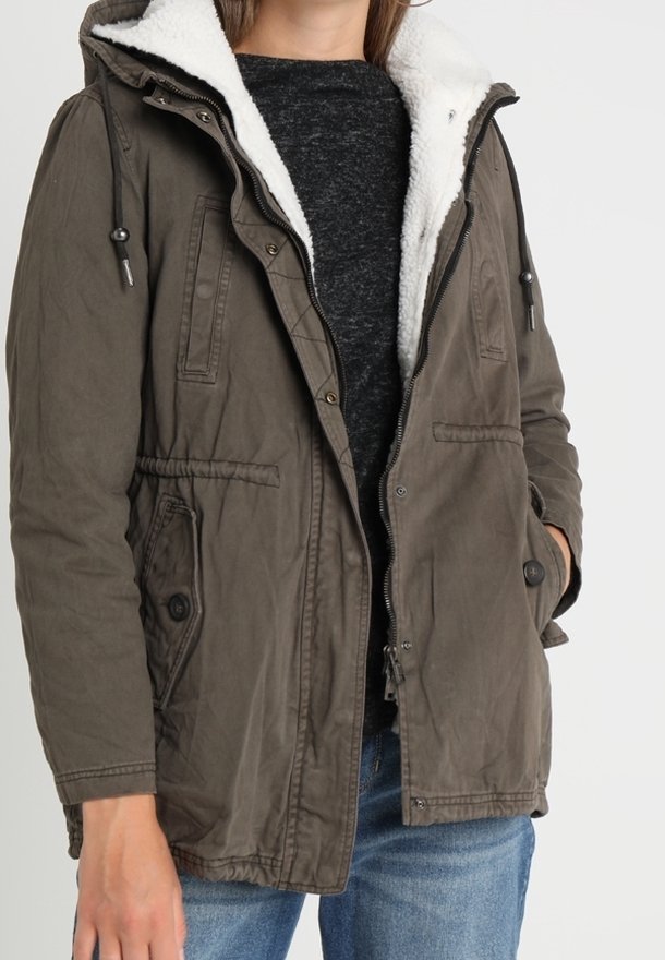 Chaqueta parka de color verde oliva con un forro de forro polar blanco esponjoso, capucha con cordón, bolsillos frontales y cierre de cremallera, llevada sobre una camisa oscura.