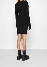 Robe noire en maille avec manches longues, dotée d'un design structuré et de liens aux poignets. Associée à des bottines noires à lacets.