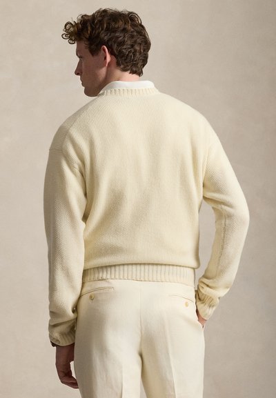 Άνδρας με σγουρά μαλλιά γυρισμένος προς τα πίσω, φοράει κρεμ πουλόβερ πάνω από λευκό πουκάμισο με γιακά και κρεμ παντελόνι.